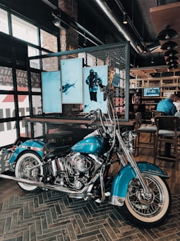 A blue motorcycle with chrome details is parked indoors on a checkered floor. Behind it, a screen displays images of a scuba diver and a shark in an aquatic setting. The room has exposed brick walls and industrial ceiling lights.