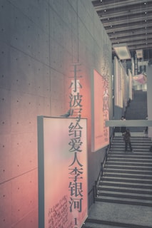 A modern interior space with a large concrete wall adorned with artistic banners in Chinese characters. The setting includes a staircase and architectural elements such as beams and high ceilings. People are casually interacting and moving in this urban environment, suggesting a public or cultural venue.