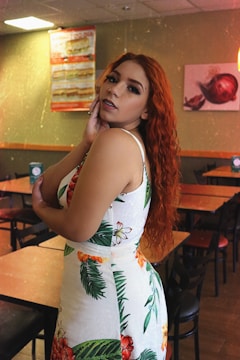 A woman with long curly red hair stands in a restaurant setting, wearing a white dress with a tropical floral pattern. She poses with one hand touching her face, looking directly at the camera. The restaurant has wooden tables and chairs, with a warm ambiance enhanced by wall decorations.