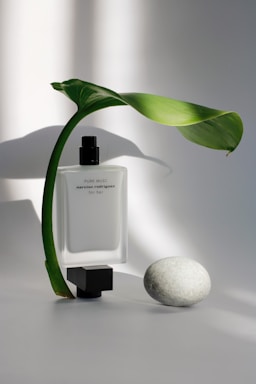 A minimalist arrangement featuring a perfume bottle with a sleek white design and black cap, labeled 'Pure Musc' by Narciso Rodriguez for her. A single green leaf elegantly arches over the bottle, and a smooth white stone rests nearby, all set against a soft, diffused light backdrop.