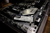 A close-up view of a computer motherboard with a Kingston A2000 NVMe solid-state drive placed on top. The motherboard features complex circuitry and components including slots, capacitors, and a heatsink labeled MSI. The SSD is prominently positioned, displaying clear labeling and specifications.