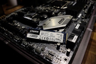 A close-up view of a computer motherboard with a Kingston A2000 NVMe solid-state drive placed on top. The motherboard features complex circuitry and components including slots, capacitors, and a heatsink labeled MSI. The SSD is prominently positioned, displaying clear labeling and specifications.