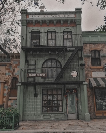 A vintage building with a green facade displaying a sign that reads 'Hudson Pawn Shop' above the entrance. The building features large windows and a fire escape ladder, while a smaller sign advertises 'Rooms for Rent'. Trees with sparse leaves are visible in the foreground, enhancing the urban setting.