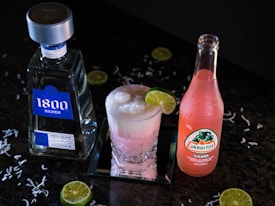 A bottle of 1800 Silver Tequila, an ice-filled crystal glass with a frothy pink drink garnished with a lime slice, and a bottle of Jarritos Guava soda are placed on a dark surface. Lime slices and white shredded pieces are scattered around.