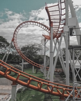 An outdoor roller coaster track featuring a series of loops and spirals, supported by white steel pillars. The track is predominantly orange and stands against a partly cloudy sky. Green trees and grass are visible in the background, adding to the park-like atmosphere.