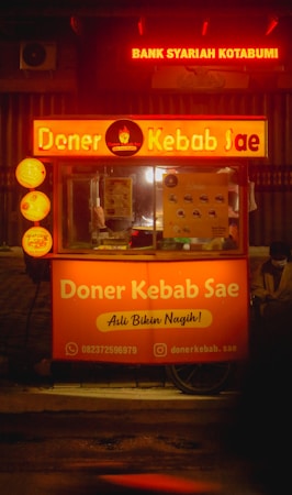 A kebab stand with bright orange signage displaying 'Doner Kebab Sae' is illuminated in a dimly lit street. The stand features contact information and social media handles. Above, red signage for 'Bank Syariah Kotabumi' is visible. A person is sitting on the right side, partially obscured by the stand.