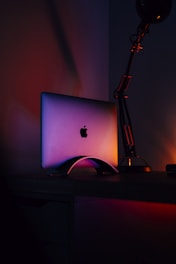 A laptop with an illuminated logo stands on a desk, surrounded by a warm glow with a dark ambient background. The light casts colorful hues on the device, creating a purple and orange gradient effect.