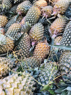 A pile of fresh pineapples with spiky green leaves and rough, textured outer skin in shades of green and yellow. The fruits are haphazardly arranged, some with more golden tones indicating ripeness, and others with varying shades of green.