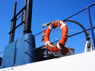 A lifebuoy with the words 'TORONTO' and 'ORIGINAL' is attached to the railing of a ship. The lifebuoy is orange and white, and it stands out against the bright blue sky. The metal structure of the ship includes ropes and a dark vertical exhaust pipe.