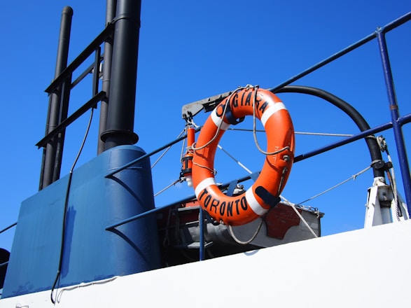 A lifebuoy with the words 'TORONTO' and 'ORIGINAL' is attached to the railing of a ship. The lifebuoy is orange and white, and it stands out against the bright blue sky. The metal structure of the ship includes ropes and a dark vertical exhaust pipe.