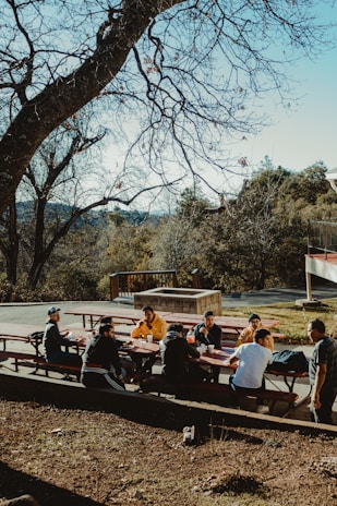 Smiling faces of people gathered around a picnic table enjoying outdoor food.