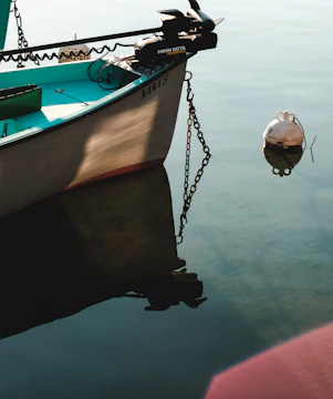 A small boat is moored in calm water, with a visible anchor chain extending to a buoy floating nearby. The boat is named, and part of its aqua-colored interior can be seen, along with a motor attached at the stern.