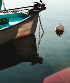 A small boat is moored in calm water, with a visible anchor chain extending to a buoy floating nearby. The boat is named, and part of its aqua-colored interior can be seen, along with a motor attached at the stern.