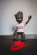 A small figure resembling a tree character is sitting on a Nike shoebox. The character is wearing a white t-shirt with a red Supreme logo. The surface is predominantly dark with a prominent red Nike logo.