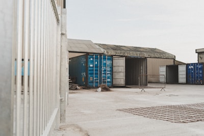 A fenced industrial area containing several shipping containers, some of which are blue and open, is situated on a concrete lot surrounded by industrial buildings.
