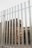 Tall, neatly stacked wooden pallets are visible behind a tall metal fence with vertical bars. The pallets are arranged in an orderly fashion and there is an industrial building visible in the background.