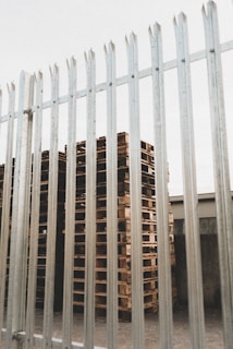 Tall, neatly stacked wooden pallets are visible behind a tall metal fence with vertical bars. The pallets are arranged in an orderly fashion and there is an industrial building visible in the background.