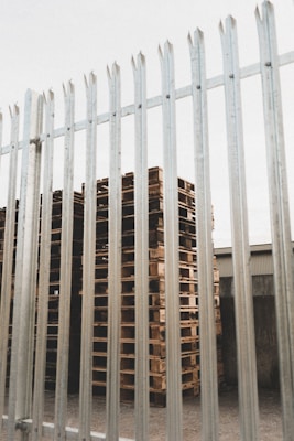 Tall, neatly stacked wooden pallets are visible behind a tall metal fence with vertical bars. The pallets are arranged in an orderly fashion and there is an industrial building visible in the background.