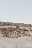 A light brown industrial building has a roof with several vents; in front of it are multiple stacks of wooden pallets and several large containers. In the foreground, there are two large piles of broken concrete and rubble.