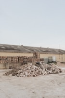 A light brown industrial building has a roof with several vents; in front of it are multiple stacks of wooden pallets and several large containers. In the foreground, there are two large piles of broken concrete and rubble.