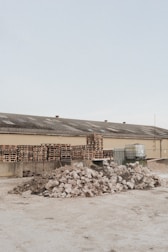A light brown industrial building has a roof with several vents; in front of it are multiple stacks of wooden pallets and several large containers. In the foreground, there are two large piles of broken concrete and rubble.