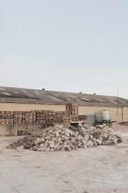 A light brown industrial building has a roof with several vents; in front of it are multiple stacks of wooden pallets and several large containers. In the foreground, there are two large piles of broken concrete and rubble.
