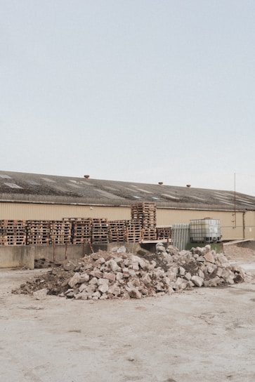 A light brown industrial building has a roof with several vents; in front of it are multiple stacks of wooden pallets and several large containers. In the foreground, there are two large piles of broken concrete and rubble.