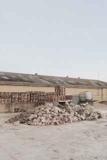 A light brown industrial building has a roof with several vents; in front of it are multiple stacks of wooden pallets and several large containers. In the foreground, there are two large piles of broken concrete and rubble.