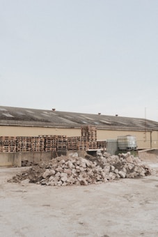 A light brown industrial building has a roof with several vents; in front of it are multiple stacks of wooden pallets and several large containers. In the foreground, there are two large piles of broken concrete and rubble.