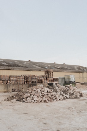 A light brown industrial building has a roof with several vents; in front of it are multiple stacks of wooden pallets and several large containers. In the foreground, there are two large piles of broken concrete and rubble.