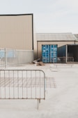 A fenced industrial area contains shipping containers and construction materials. Metal barriers are placed around, and the environment appears to be a storage or construction site.