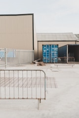 A fenced industrial area contains shipping containers and construction materials. Metal barriers are placed around, and the environment appears to be a storage or construction site.