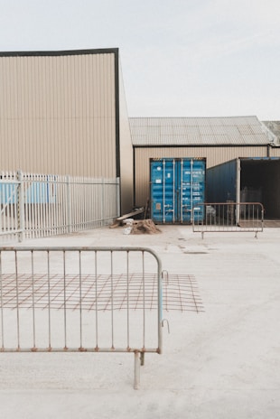 A fenced industrial area contains shipping containers and construction materials. Metal barriers are placed around, and the environment appears to be a storage or construction site.