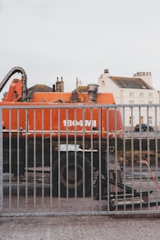 A large orange industrial vehicle with the label '1804 MI' is positioned behind a metal fence. The vehicle appears to be a construction or excavation machine, with components like pipes and a large wheel visible. In the background, there are residential buildings with multiple windows and chimneys, suggesting an urban setting.