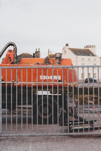 A large orange industrial vehicle with the label '1804 MI' is positioned behind a metal fence. The vehicle appears to be a construction or excavation machine, with components like pipes and a large wheel visible. In the background, there are residential buildings with multiple windows and chimneys, suggesting an urban setting.