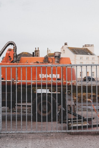 A large orange industrial vehicle with the label '1804 MI' is positioned behind a metal fence. The vehicle appears to be a construction or excavation machine, with components like pipes and a large wheel visible. In the background, there are residential buildings with multiple windows and chimneys, suggesting an urban setting.