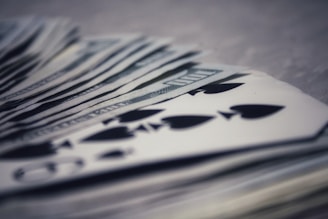 A stack of playing cards, primarily focusing on the spade suit, is overlaid on top of a bundle of US dollar bills. The image blurs slightly in the background, highlighting foreground details and creating a sense of depth.