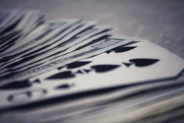 A stack of playing cards, primarily focusing on the spade suit, is overlaid on top of a bundle of US dollar bills. The image blurs slightly in the background, highlighting foreground details and creating a sense of depth.