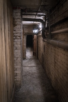 A dimly lit, narrow corridor with brick walls and a concrete floor. The space features exposed pipes and a single light bulb illuminating part of the hallway. An open doorway leads into a darker area, creating an air of mystery and suspense.