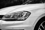 Close-up view of the front left headlight of a modern car, displaying the sleek and smooth curves of the vehicle's design. The focus on the headlight highlights the precision in the car's craftsmanship, with reflections visible on the shiny surface of the white paint.