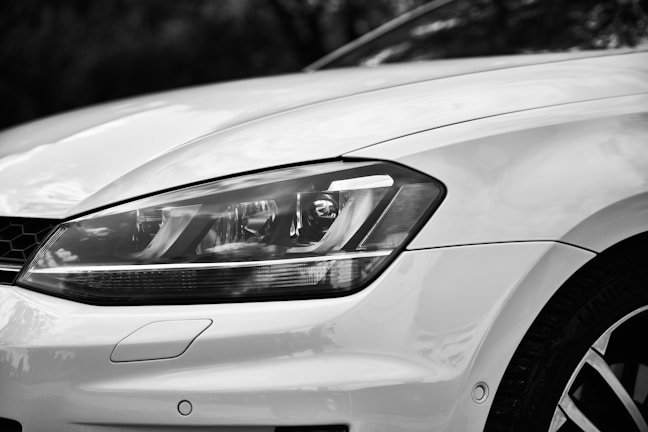 Close-up view of the front left headlight of a modern car, displaying the sleek and smooth curves of the vehicle's design. The focus on the headlight highlights the precision in the car's craftsmanship, with reflections visible on the shiny surface of the white paint.