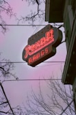 A neon sign for a Rexall drugstore is prominently displayed against a dusky sky. The letters are glowing in bright red, and the sign appears slightly vintage. Bare tree branches frame the scene, adding a stark contrast to the glowing sign.