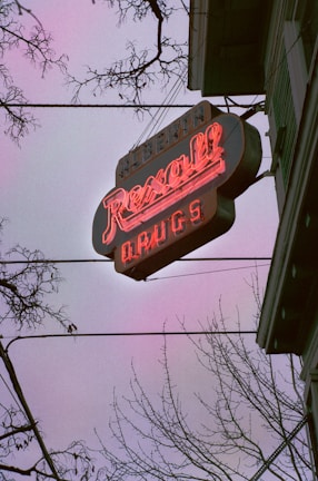A neon sign for a Rexall drugstore is prominently displayed against a dusky sky. The letters are glowing in bright red, and the sign appears slightly vintage. Bare tree branches frame the scene, adding a stark contrast to the glowing sign.