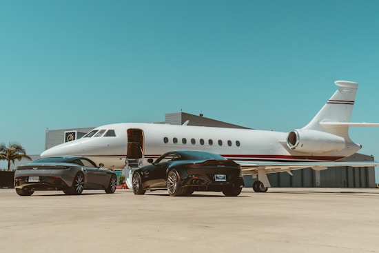 A sleek private jet is parked on an airport tarmac near two luxury black sports cars. The jet's staircase is extended, suggesting boarding or deboarding. The setting is clear and sunny, with a blue sky in the background and hangars nearby.
