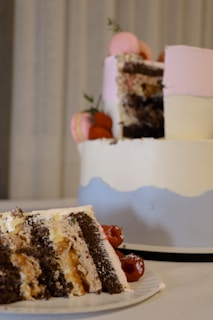 A layered cake with pink and white frosting, partially eaten, is decorated with macarons and strawberries. In the foreground, a slice of the cake showing the layers of chocolate and vanilla as well as a filling, likely fruit or cream, is placed on a white plate.