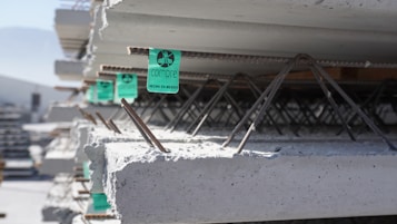 Stacks of concrete slabs are seen lined up in an orderly manner. Each slab has visible metal rebars embedded within, and some have green labels attached that read 'Compre, Hecho en M&eacute;xico'. Sunlight casts shadows on the concrete, suggesting it is taken in an outdoor industrial setting.