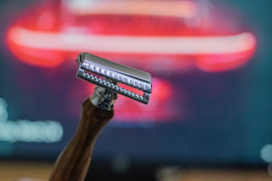 A close-up of a safety razor with a wooden handle, set against a blurred background with red and black hues. The razor is in sharp focus, emphasizing its metallic shine and the texture of the handle.