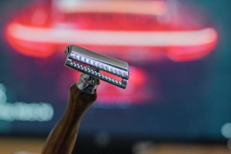 A close-up of a safety razor with a wooden handle, set against a blurred background with red and black hues. The razor is in sharp focus, emphasizing its metallic shine and the texture of the handle.