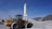 A large yellow wheel loader with the brand 'CASE' written on its side is positioned on a construction site, next to a pile of gray gravel. In the background, there is a tall cylindrical structure with the word 'Cemex' written on it. The sky is clear and blue, with mountains visible in the distance.