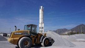 A large yellow wheel loader with the brand 'CASE' written on its side is positioned on a construction site, next to a pile of gray gravel. In the background, there is a tall cylindrical structure with the word 'Cemex' written on it. The sky is clear and blue, with mountains visible in the distance.